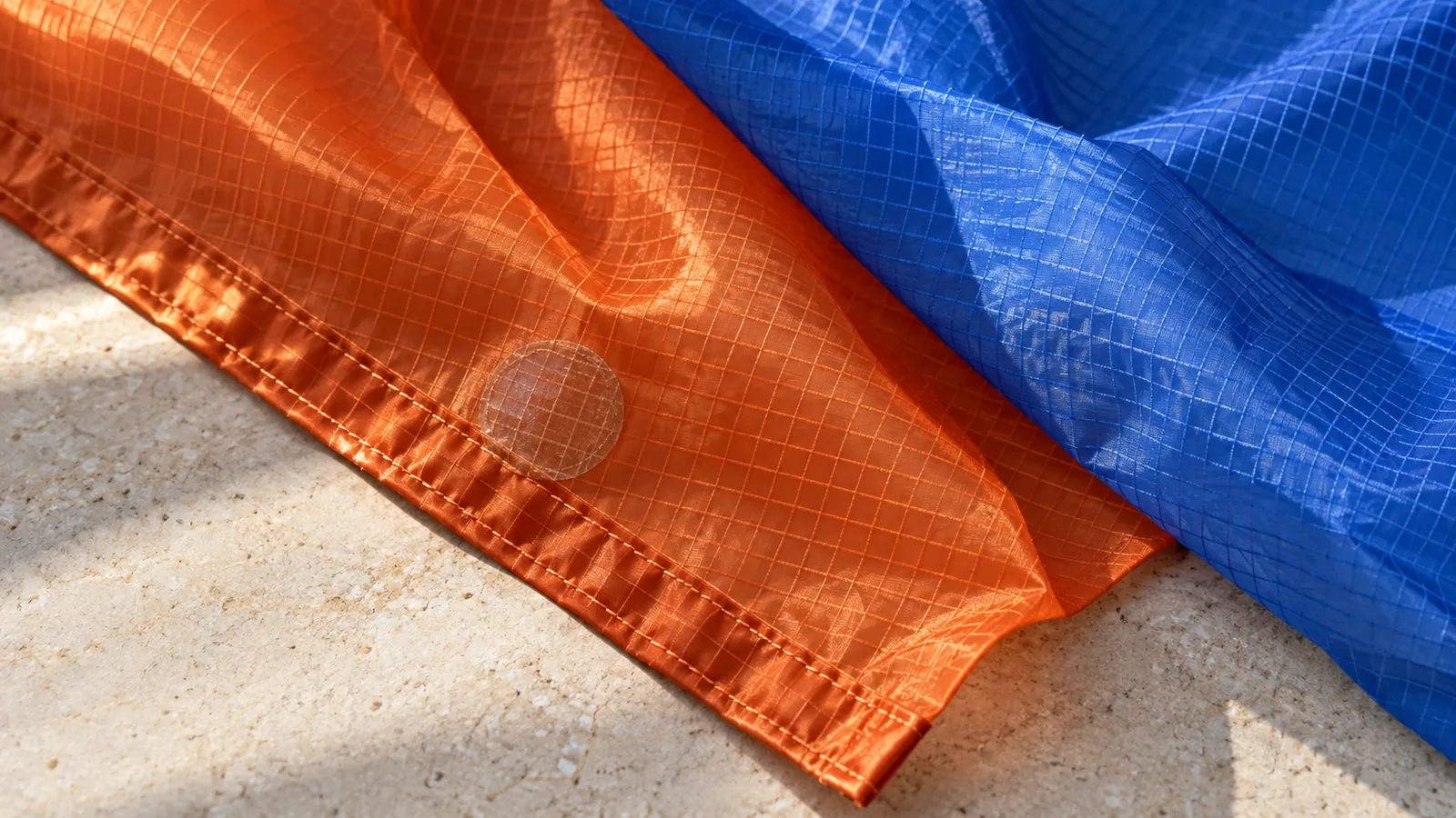 Illustration: close-up of orange and blue ripstop kite fabric showing the square reinforcement grid, a stitched hem and a clear round repair patch
