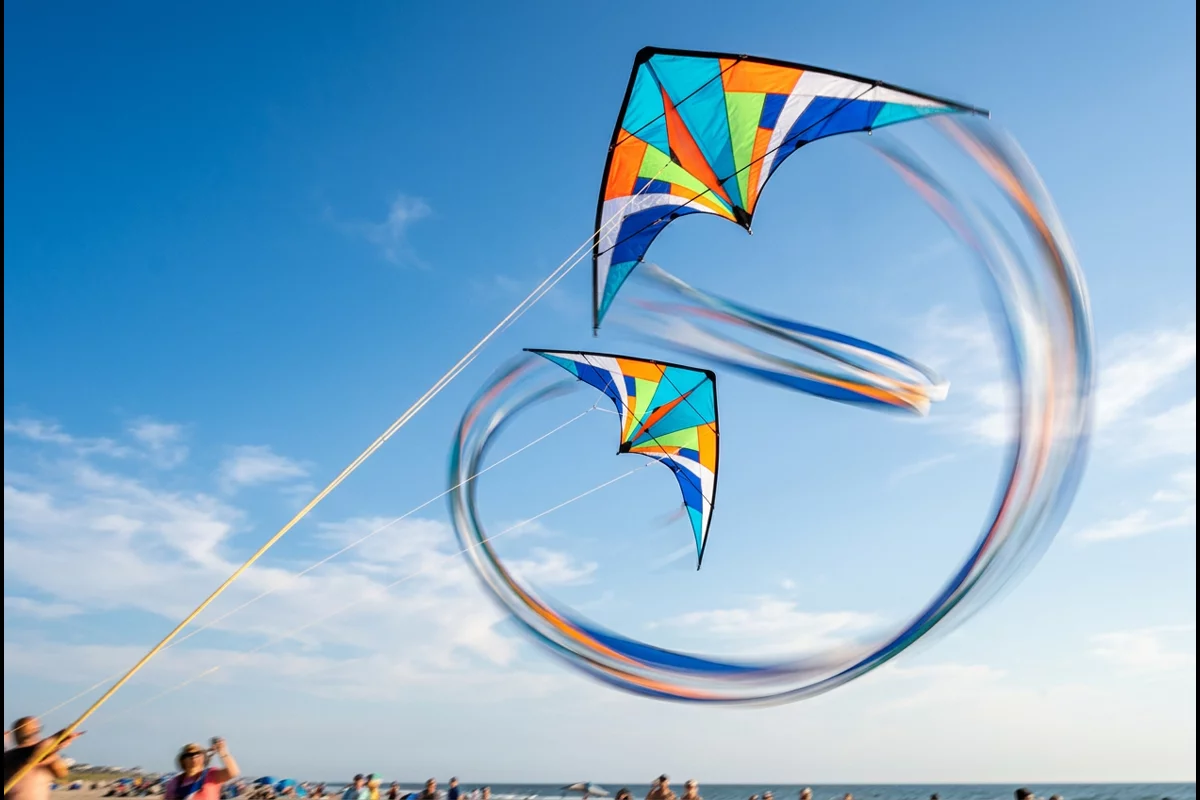 Two dual-line sport kites carving synchronized tight loops in blue sky above beach